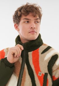 Chaqueta de forro polar con cuello verde y cremallera frontal, que presenta patrones en naranja, beige y verde oscuro. Incluye un pequeño logo en el lado izquierdo del pecho.