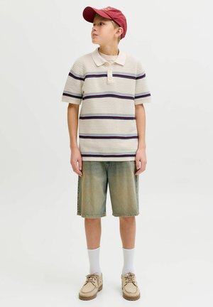 Berniukas dėvi raudoną kepurę, dryžuotą trumpomis rankovėmis polo marškinėlį, žalsvus džinsinius šortus, baltas kojines ir smėlio spalvos batus, žiūri į šoną.