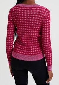 Stickad långärmad tröja i rosa med ett rutmönster. Har ribbade fållar och en rund halsringning, och visar upp en strukturerad tygdesign.