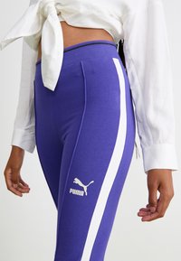 Lila sportleggings med vita sidostråk, med en ribbad midja och en vit Puma-logotyp på underbenet. Slät textur.