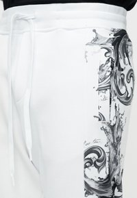 Λευκή φόρμα με πλαϊνές τσέπες, διακοσμημένη με μοτίβο μαύρων και γκρι αφηρημένων στροβίλων στη μία πλευρά και λευκή κορδόνι στη μέση.