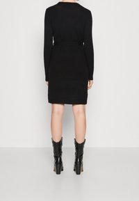 Abito maglione nero con maniche lunghe, texture del tessuto visibile. Include una cintura abbinata, lunghezza al ginocchio, abbinato a stivali neri con tacco.