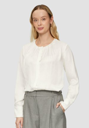 Witte blouse met lange mouwen, ronde hals en geplooide details bij de schouders. De stof lijkt glad en zijden, gecombineerd met grijze broeken.