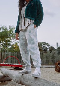 Turkusowa polarowa kurtka, biała koszulka w tie-dye, jasnoszare dresy i białe trampki z szarymi akcentami; model stoi na kłodzie w placu zabaw.
