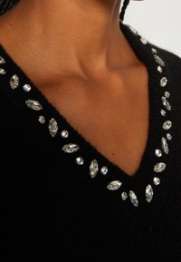 Pull noir à col en V orné de strass clairs taillés en rond et en marquise alternés le long de l'encolure sur une peau brune.