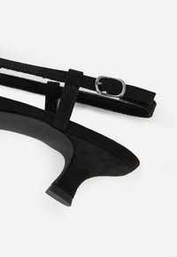 Zapato de tacón alto de ante negro con punta afilada y tacón estrecho. Incluye una correa negra ajustable con un detalle de hebilla plateada.