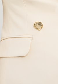 Gros plan sur un blazer beige crème ajusté avec une poche à rabat et un bouton doré brillant de forme irrégulière.