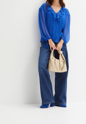 Femme en blouse bleue à volants et jean à jambes larges tenant un sac à main beige, debout sur un fond blanc.