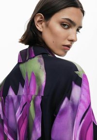 Camicia fluida con un design floreale caratterizzato da grandi elementi viola e verdi su uno sfondo scuro. Texture liscia con un colletto strutturato.