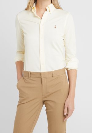 Kobieta ubrana w jasnożółtą koszulę na guziki z podwiniętymi rękawami i beżowe spodnie, z jedną ręką w kieszeni, stoi na jednolitym tle.