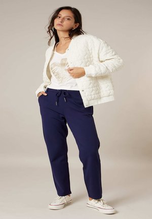 Femme portant une veste matelassée blanche sur une chemise blanche avec un motif doré, un pantalon à cordon de serrage bleu marine et des baskets blanches, debout devant un fond beige.