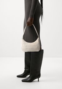 Kremowa torebka o prążkowanej teksturze, zakrzywionym kształcie i pojedynczym pasku. Połączona z wysokimi, czarnymi skórzanymi botkami.