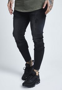 Svarta slitna skinny jeans med flera hål och urtvättning. Matchas med svarta sneakers, som har en slät yta och texturerade detaljer.