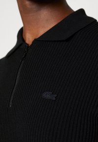 Polo noir à côtes avec un col zippé, présentant un logo bleu marine brodé sur la poitrine gauche. Détail de tissu texturé visible.