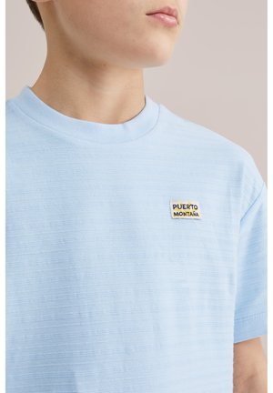 Młoda osoba ubrana w jasnoniebieską teksturowaną koszulkę z małą przypinką "Puerto Montana" na lewej piersi, neutralne tło.