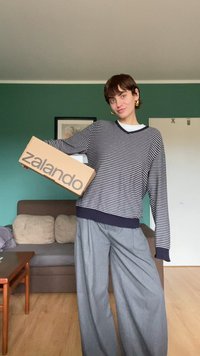 Juostuotas tamsiai mėlynas megztinis su V formos kaklu, derinamas su plačiomis pilkų spalvos kelnėmis; laiko šviesiai rudos spalvos „Zalando“ dėžutę. Minkštos tekstūros, kasdienis dizainas.