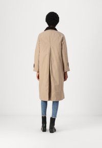 Beige trenches kabát sötét színű gallérral, hosszú ujjakkal és hátsó redőzéssel, kék farmerral és fekete bokacsizmával párosítva.