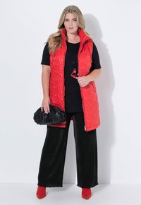 Rote gesteppte Weste mit hohem Kragen über einem schwarzen T-Shirt mit Lippen-Print. Schwarze, plissierte, weit geschnittene Hosen und rote Ankle Boots vervollständigen das Outfit.
