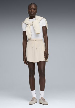 Dunkelhäutige Person steht, trägt ein weißes T-Shirt, einen beigen Rock und einen Pullover über den Schultern, dazu weiße Socken und beige Sneakers.