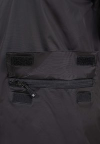 Tissu noir avec une poche horizontale à fermeture éclair sécurisée par deux bandes Velcro au-dessus et en dessous de la fermeture éclair.