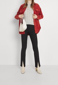 Chemise oversized en velours côtelé rouge, pull en maille câble gris clair, pantalons noirs avec fentes sur les côtés, et bottines à talons beiges, accompagnés d'un sac blanc.