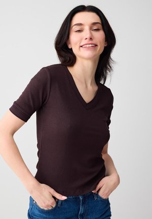 Giovane donna sorridente, che indossa una maglietta a maniche corte color marrone scuro con scollo a V a coste e jeans blu, con le mani in tasca, su sfondo semplice.