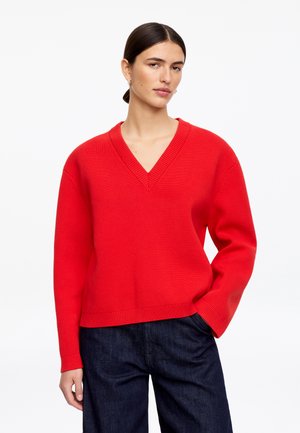 Kobieta ubrana w jasnoczerwony sweter z dekoltem w serek i ciemnoniebieskie dżinsy, stojąca z jedną ręką w kieszeni na jednolitym białym tle.