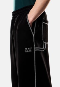 Pantaloni da tuta neri con vita elastica, cuciture bianche a contrasto, tasche laterali e logo EA7. Tessuto morbido con vestibilità rilassata.