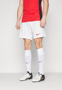 Rot und schwarz kurzärmliges Hemd, weiße Shorts mit rotem Nike-Logo, weiße Socken mit blauem Nike-Logo, schwarz-weiße Fußballschuhe.