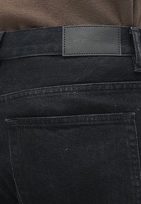 Pantalones de mezclilla negra con una etiqueta de cuero con la marca "SAMSOE SAMSOE", visible en la cintura. La tela tiene una textura mate.