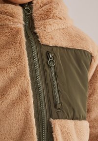 Chaqueta de pelaje sintético marrón con un panel de nailon verde olivo y cremallera, que incluye un cuello alto y un bolsillo lateral para almacenamiento.