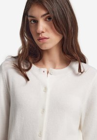 Vit stickad cardigan med rundad halsringning, prydd med en rad av fyra knappar. Materialen verkar mjuka med en slät textur.