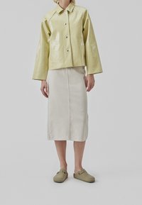 Hellgrüne, krokodilledergeprägte Jacke mit kragenlosem Hals, kombiniert mit einem cremefarbenen, knielangen Rock und olivgrünen Holzpantoffeln.