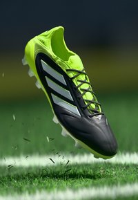 Soccer cleat in neon yellow and black with white stripes, suspended mid-air above grass with small dirt and grass particles.