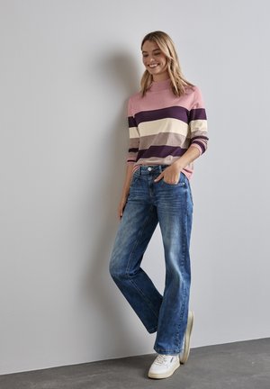 Gestreifter Pullover in Rosa-, Creme- und lila Tönen, kombiniert mit blauen Jeans. Das Model trägt weiße Sneakers. Neutraler Hintergrund.