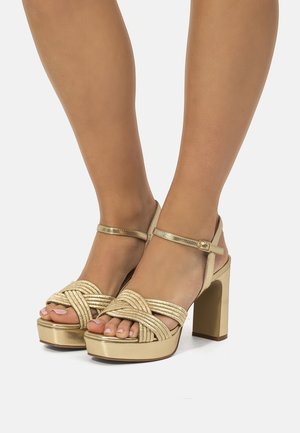 Pies con sandalias doradas de plataforma y tacón alto con tiras trenzadas y hebillas en el tobillo sobre un fondo blanco.