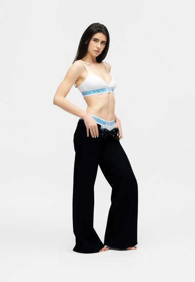 Γυναίκα με λευκό crop top και εσώρουχο με μπλε ελληνικό κλειδί, τραβάει προς τα κάτω μαύρο παντελόνι φαρδιάς γραμμής, στέκεται μπροστά σε απλό φόντο.