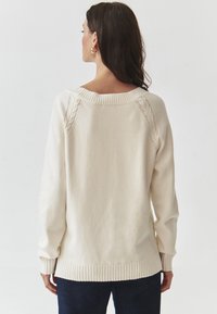 Pulover de culoare crem, cu un design din tricotaj cu cabluri pe umeri, cu manșete și tiv cu dungi. Textură clară și croială lejeră.