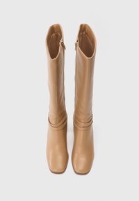 Bottes en cuir tan, allant jusqu'au genou, avec un bout carré, équipées d'une fermeture éclair sur le côté et d'un détail de sangle enroulée près de la cheville. Texture lisse.