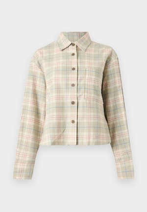 Dark-skinned woman stands with hand on hip wearing beige plaid buttoned shirt over white top, baggy blue jeans, and navy sneakers.