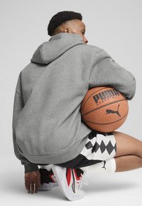 Un sweat à capuche gris, un short à motifs noir et blanc, et des baskets rouge-noir-blanc. Tenant un ballon de basket avec le logo "PUMA BASKETBALL" visible.