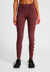 Bordeauxrode legging met een gladde textuur, hoge tailleband en witte chevron-accenten aan de zijkant. Gecombineerd met zwarte sportschoenen.