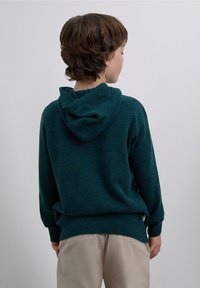 Mörkgrön stickad hoodie med huva, ribbade muddar och fåll samt en något lös passform. Sedd bakifrån med en mjuk textur.