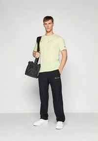 Svetlo zelena majica s vezenim logotipom, črne športne hlače z logotipom in beli supergi. Model drži črno torbico.