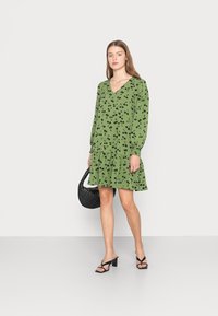 Vestido floral verde com padrões pretos, decote em V, mangas longas, barra com babados, combinado com uma mala preta e sapatos de salto abertos pretos.