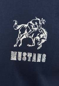 Rider wearing a hat rides a bucking horse above the word "MUSTANG" in bold, textured font on dark fabric background.