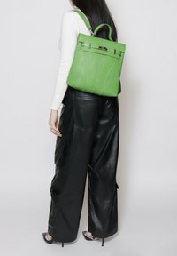Groene leren rugzak met handvat bovenaan, gladde textuur en zilveren hardware accenten. Geselecteerd bij een witte outfit en zwarte leren broek.