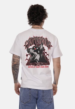 Uomo con capelli ricci e scuri che indossa una maglietta bianca con la grafica di un cane nero aggressivo in una figura triangolare a catena e il testo "PROPAGANDA ENTRATE A VOSTRO RISCHIO."