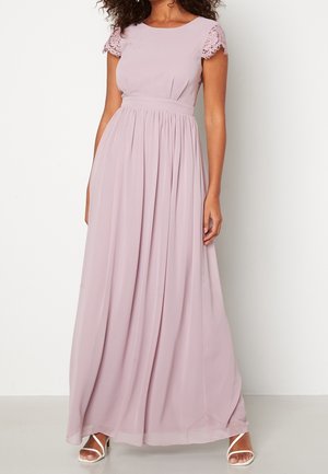 Frau in einem bodenlangen Lavendelfarbenen Kleid mit Spitzenärmeln und plissiertem Rock, kombiniert mit weißen, hochhackigen Sandalen mit Riemchen.