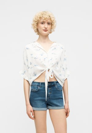 Jonge vrouw met kort krullend blond haar, gekleed in een wit bloemenoverhemd met strik aan de voorkant en hooggetailleerde opgerolde denim shorts, staand tegen een effen achtergrond.